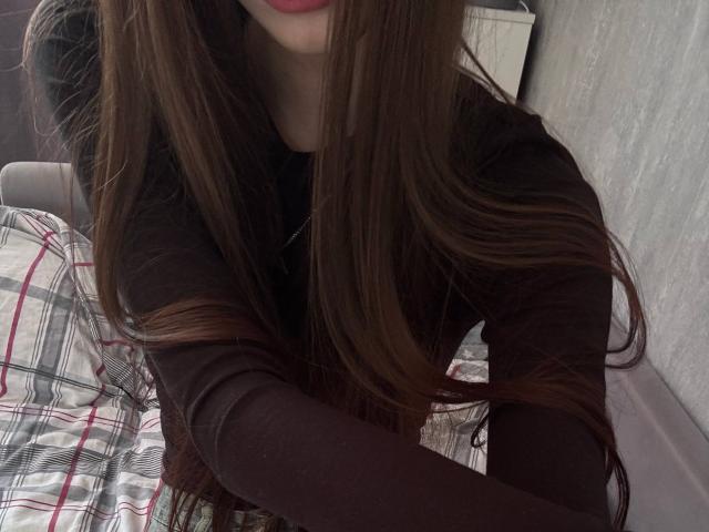 CatinaLetchworth with long brown hair, wearing a dark brown long-sleeve shirt, showing red lipstick, sitting on a bed with plaid bedding, in a slightly blurry selfie.