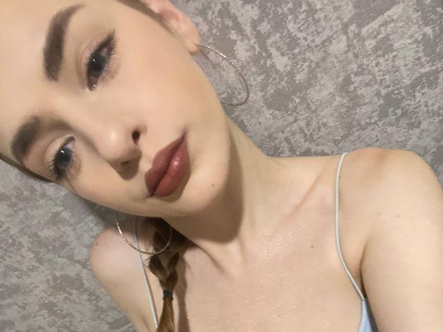 MarcelleMantz with pale skin, dark eyebrows, and red lipstick, wearing a light blue spaghetti strap top, hoop earrings, and brown braid, looking slightly down. Her tits are small.