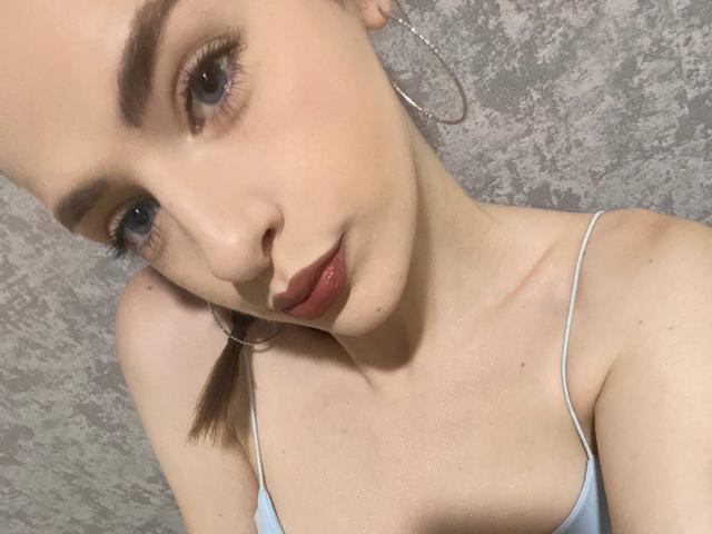 MarcelleMantz's face with light skin, brown eyebrows, blue eyes, red lipstick, small hoop earrings, and a light blue spaghetti strap top, showing her tits and a slight neck.