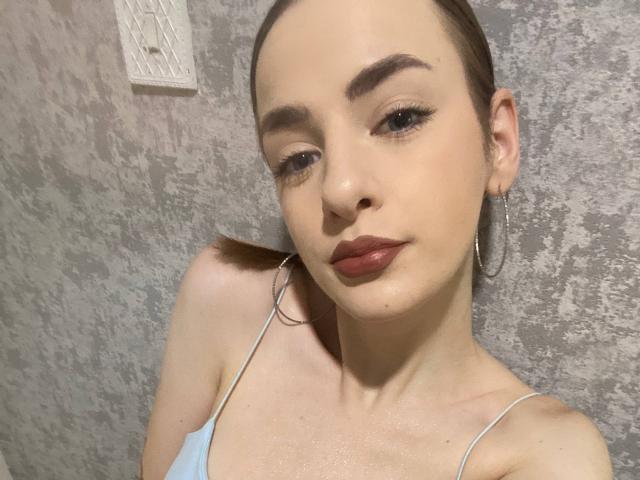 MarcelleMantz with a bald head, light skin, brown eyebrows, wearing red lipstick, hoop earrings, and a light blue spaghetti strap top, looking at the camera with a slight smile.