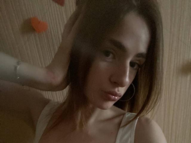 MarcelleMantz with brown hair, wearing a white top, has a hand in her hair, showing pouty lips, hoop earrings, and a slightly blurred face in dim lighting.