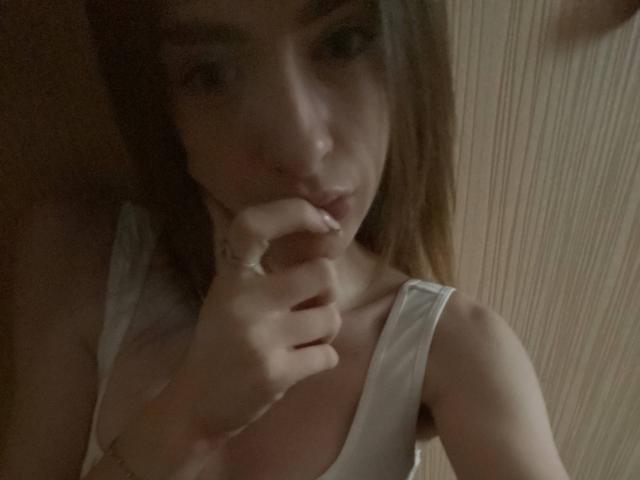 MarcelleMantz with brown hair, white tank top, and finger in mouth, showing her tits slightly. Dim lighting, focused on her face and chest.