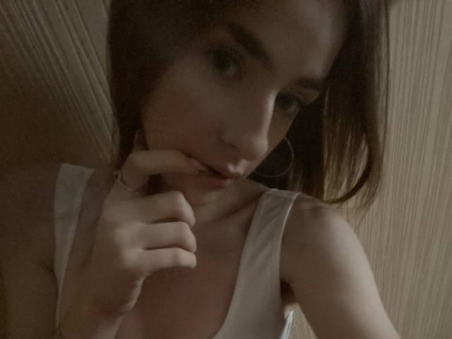 MarcelleMantz with brown hair, white tank top, and hoop earrings, biting her finger, showing her tits, in a dimly lit, wooden room.