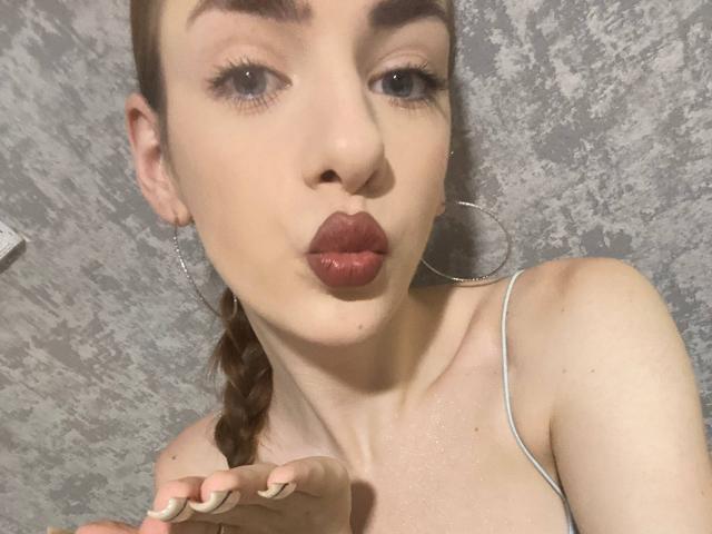MarcelleMantz with pouty lips, wearing hoop earrings, has a braided brown ponytail, light skin, and is making a kissing face, showing off her tits in a gray-patterned background.