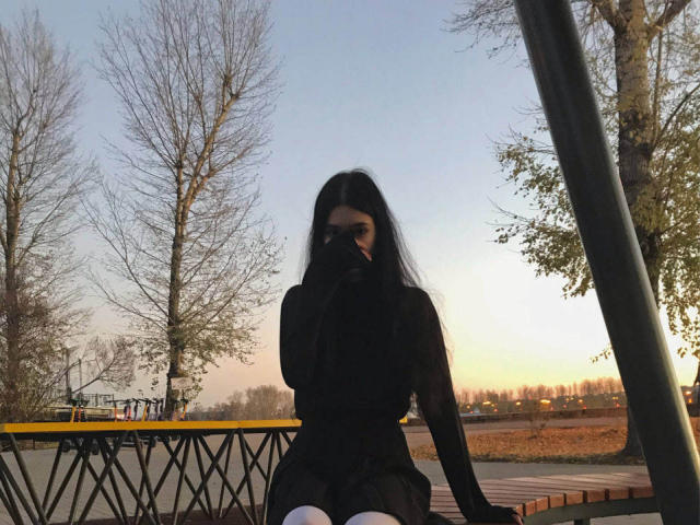 CatinaLetchworth, with long black hair, wearing a sheer black top, covering her mouth with her hand, sitting on a wooden bench, in a sunset, outside, with trees.