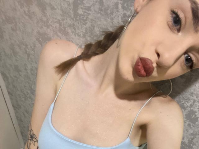 MarcelleMantz with pouty red lips, light blue spaghetti strap top, braided brown hair, hoop earrings, tattoo on right arm, and slightly glistening skin.