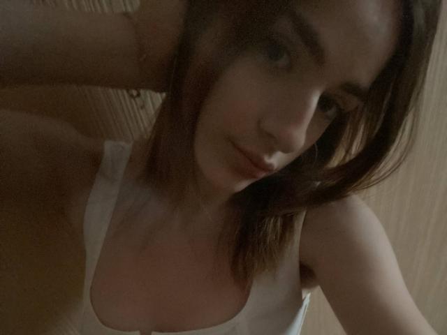 MarcelleMantz with brown hair, white tank top, and visible tits, leaning forward, looking at the camera, subtle smile, dim lighting, soft shadows on her face and chest.
