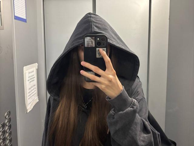 CatinaLetchworth with long brown hair, wearing a black hoodie with hood up, taking a mirror selfie in an elevator, phone covering her face, grey walls in background.
