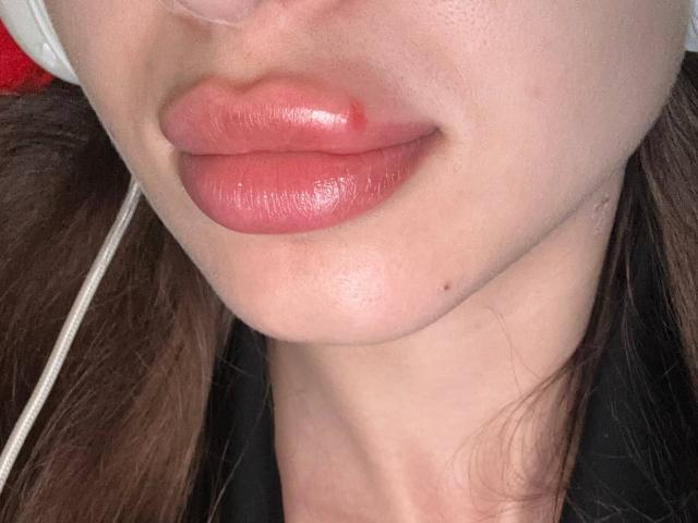 CatinaLetchworth's glossy, pinkish-red lips are slightly parted, showing a small beauty mark on her chin. White earbuds are visible on the left side of her face.