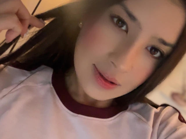 EmmaLombardi with long brown hair, pinkish skin, and brown eyes, wearing a pink and red shirt, smiling with slightly parted pink lips, holding hair on the left side.