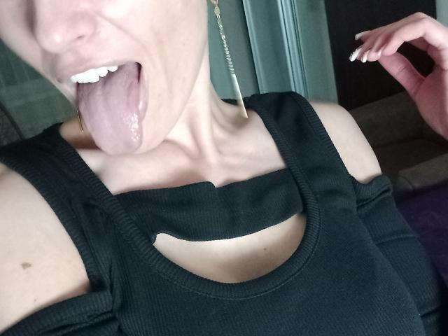 BellaJulls with tongue out, black ribbed tank top, visible cleavage, earrings, light skin, white teeth, and manicured nails, holding a straw near her mouth.