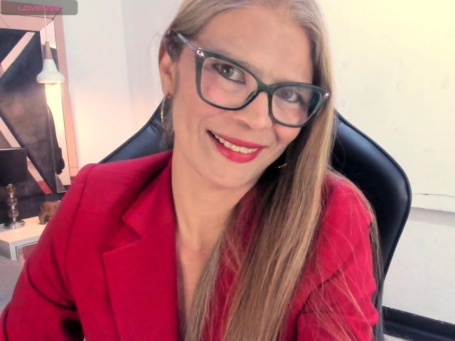 ParisMaturre with long blonde hair, green glasses, red lipstick, and red blazer, smiling at the camera, showing off her tits in a sexy, professional look.