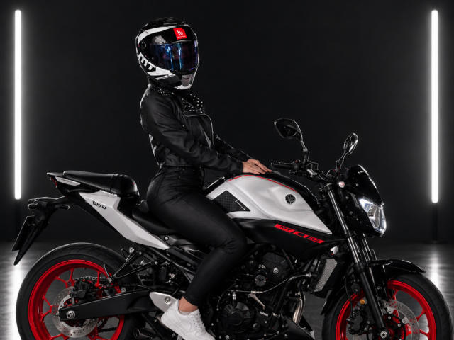 KimmyK with black leather jacket, black pants, white sneakers, and helmet, sitting on a black and white Husqvarna motorcycle with red wheels, hands on the handlebars.