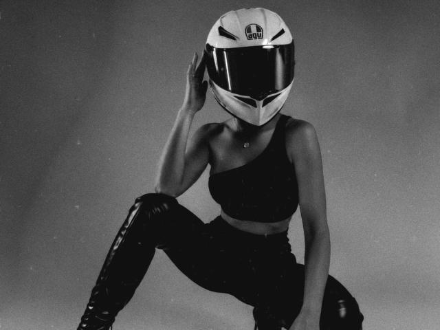 KimmyK in a black one-shoulder top and shiny black boots, squatting with a white helmet covering her face, hand touching the helmet. Greyscale, gritty.