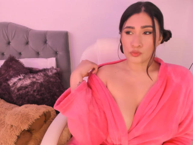 ElyRoss, Asian woman with black hair in a bun, wearing a pink robe, pulling it down to expose her small tits, sitting in a chair with a pouty expression.