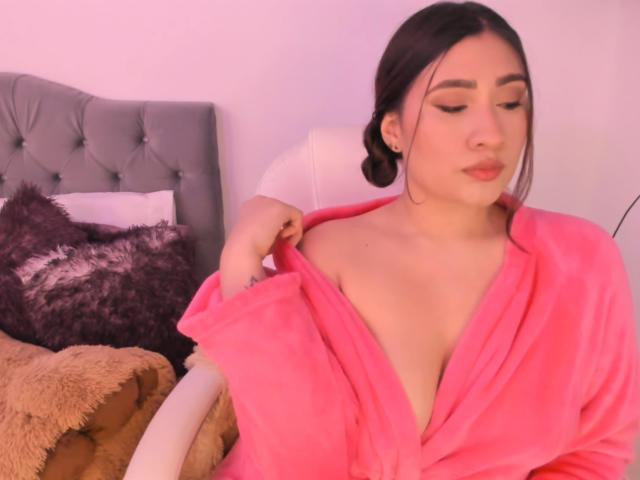 ElyRoss in a pink robe, pulling it down to reveal a modest cleavage, with dark hair in a bun, sitting on a bed with fluffy pillows.