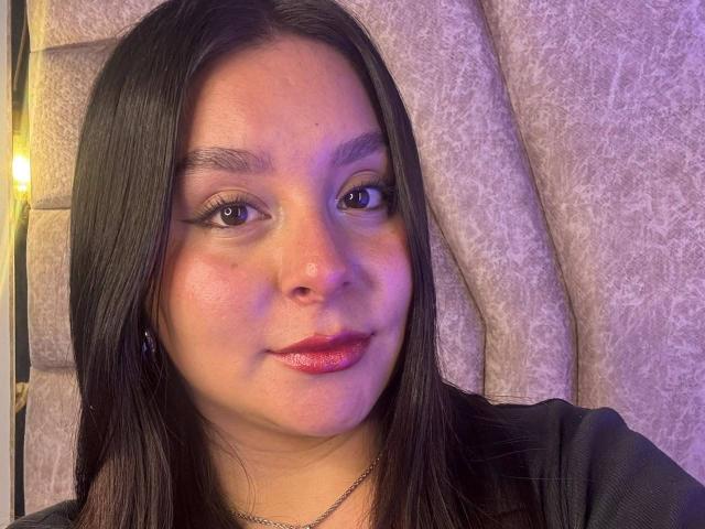 EmaJames has dark brown straight hair, medium skin, brown eyes, black eyeliner, pink lipstick, and a slight smile. She's wearing a black top and a silver necklace.