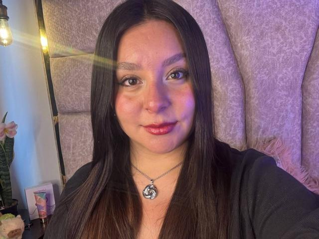 EmaJames has long brown hair, light skin, and red lipstick, wearing a black top with a silver necklace. She's smiling with a slight blush and soft purple lighting.