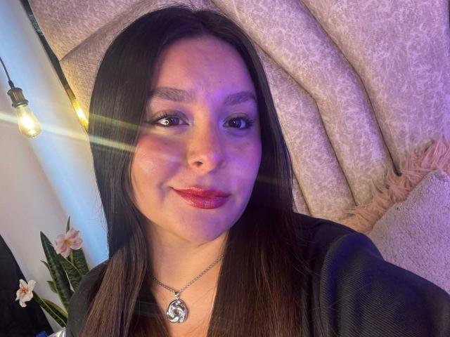 EmaJames has long brown hair, purple-tinted skin, red lipstick, and a silver necklace with a heart-shaped pendant. She's smiling with a light reflecting on her face.
