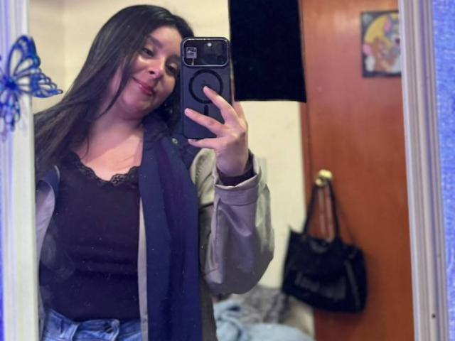 EmaJames with long brown hair, smiling, wearing a black lace top and blue jeans, taking a mirror selfie with her phone. Her tits are small, and she's wearing a jacket.