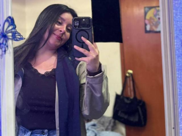 EmaJames with long brown hair, wearing a black lace top and blue jeans, taking a mirror selfie with a smug expression, showing her tits and holding a black bag.