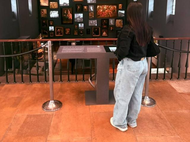EmaJames has a big ass in loose blue jeans and a black top, standing in front of a wall of framed pictures in a museum, with metal railings.