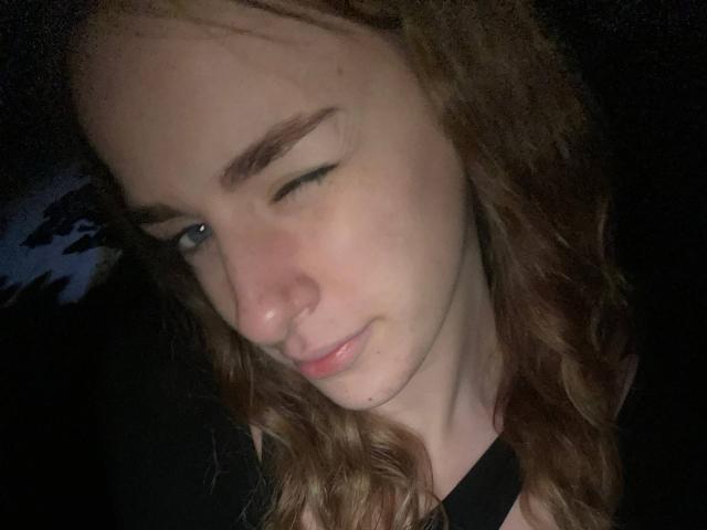 MarcelleMantz's face with winking eye, slightly flushed cheeks, and curly brown hair, wearing a black top, in a dark, dimly-lit setting. Her nose is small and cute.