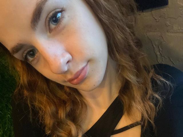 MarcelleMantz's face, showing blue eyes, wavy brown hair, and a slight smile, wearing a black top with a crisscross design, her nose is slightly upturned.