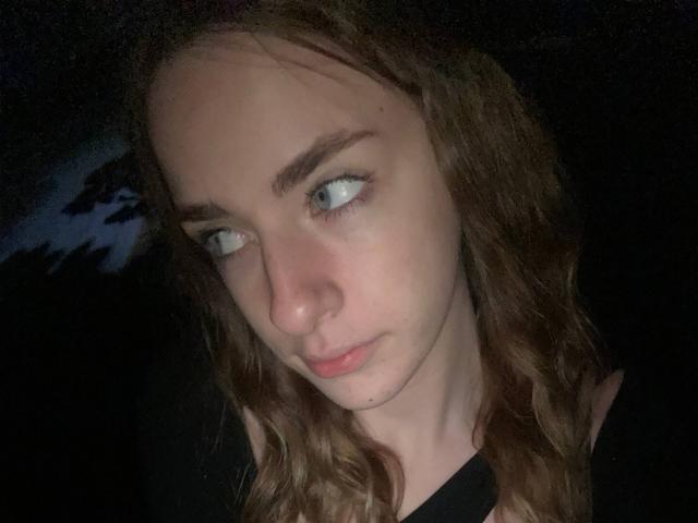 MarcelleMantz has light brown wavy hair, blue eyes, and a slight pout, looking to the side with a soft, almost sad expression in dim lighting.