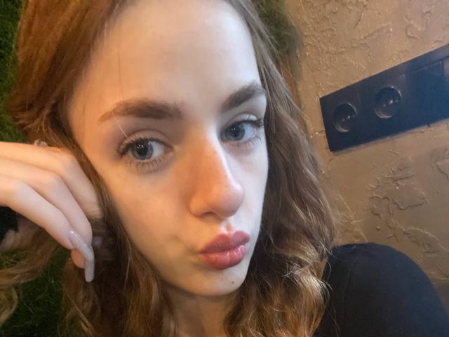MarcelleMantz has wavy brown hair, light skin, and full pink lips, pouting slightly. She's wearing a black top, with her hand near her head, blue wall behind.