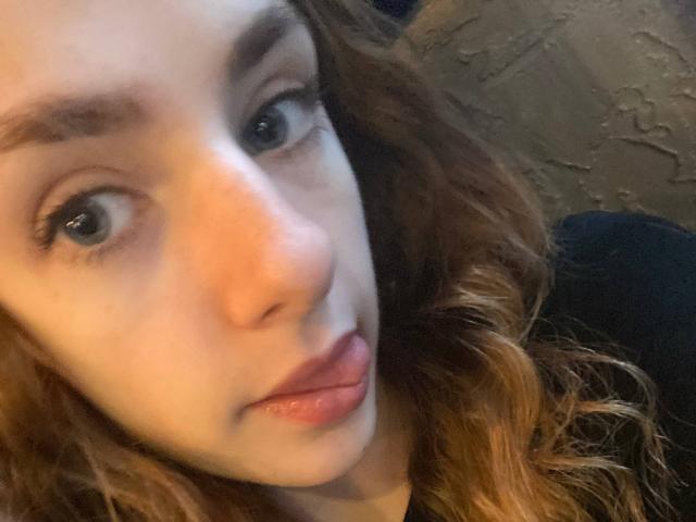 MarcelleMantz's face, with wavy brown hair, blue eyes, and slightly parted pink lips, in a tight selfie. Her eyebrows are dark and slightly raised.
