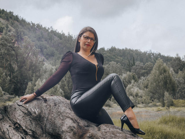 ParisMaturre, with medium tits, black leather pants, and black high heels, sits on a rock, wearing a zip-up black top and glasses, in a forested outdoor setting.
