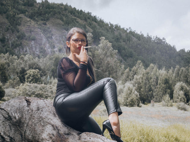 ParisMaturre with gray hair, glasses, black sheer top, and black leather pants, smoking a cigarette, sitting on a rock, wearing black heels, in a green forested mountain area.