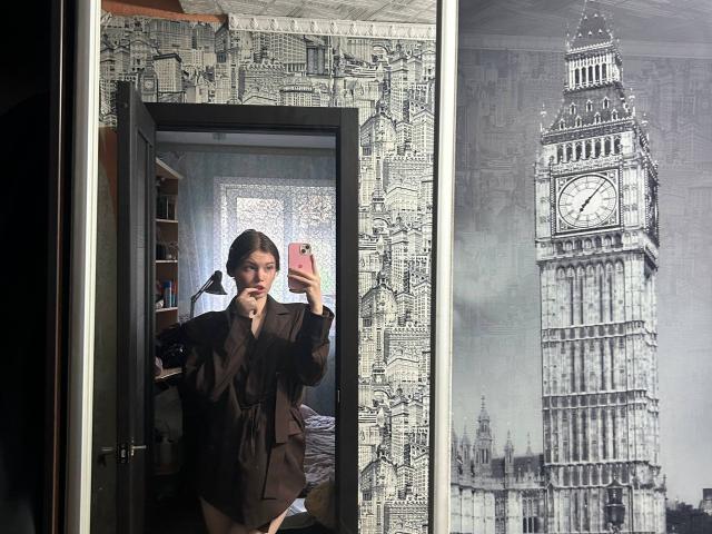 ApoloniaDavick stands in front of a mirror, taking a selfie in a brown suit, with a black clock tower on the right, holding a pink phone.