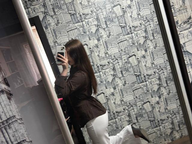 ApoloniaDavick in a brown pinstriped top and white pants, taking a mirror selfie with her phone, showing her ass and long brown hair.