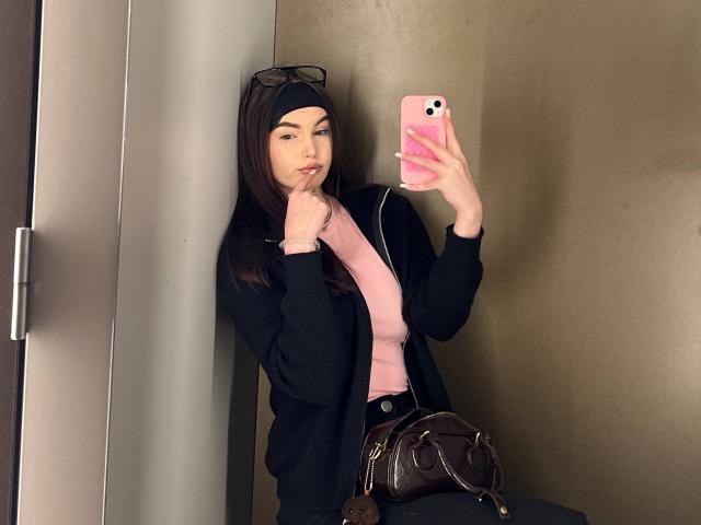 ApoloniaDavick with long brown hair, black headscarf, black jacket, pink top, holding pink phone, brown bag on knee, finger on lips, standing in corner, wearing black glasses.