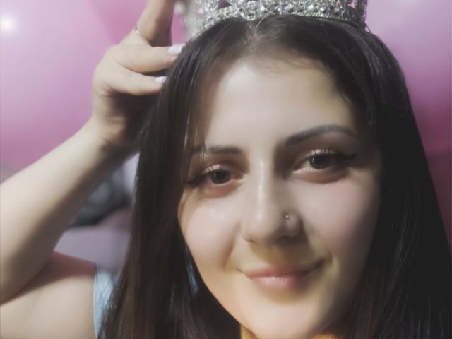 PattyAna has dark brown hair, wearing a glittery crown, smiling with a nose ring, and holding the crown with her hand, showing off her cute face and full tits.