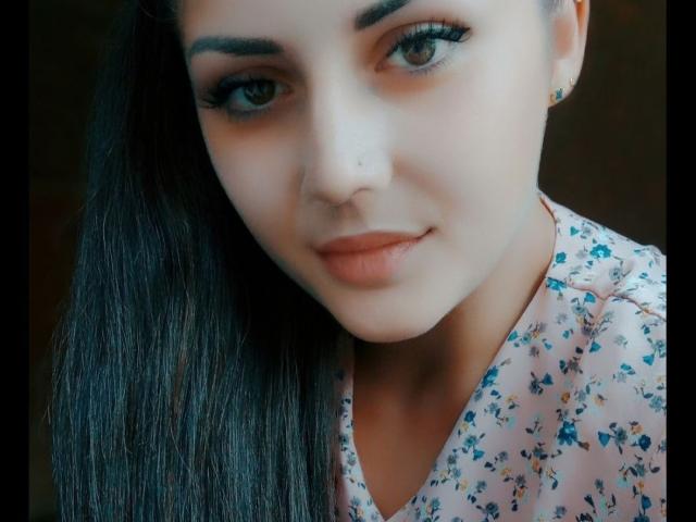 PattyAna has pale skin, dark eyebrows, long black hair, and is wearing a floral shirt with a subtle smile. Her eyelashes are thick, and she's wearing small gold earrings.
