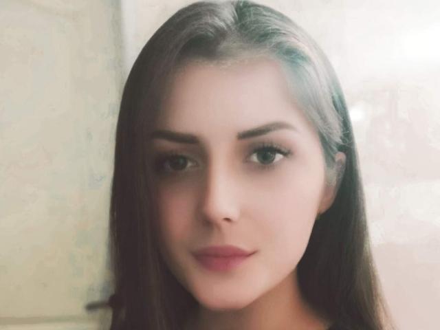 PattyAna ha lunghi capelli castani, occhi verdi e rossetto rosa. Guarda di lato con un leggero sorriso calmo. Il suo viso è centrato, morbido e leggermente sfocato..