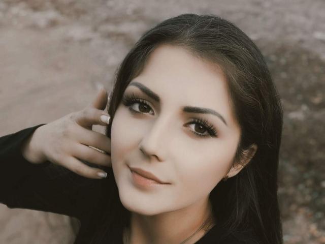 PattyAna tiene el cabello largo y oscuro, ojos marrones y labios carnosos con un maquillaje sutil. Está tocándose la cabeza con una mano, lleva una blusa negra y está mirando a la cámara..