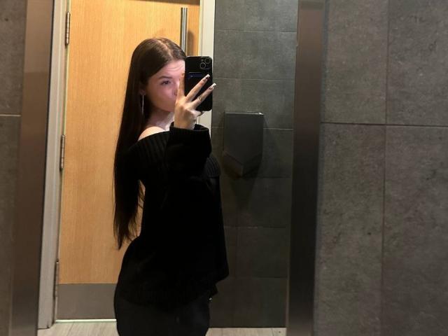 ApoloniaDavick with long brown hair, wearing an off-shoulder black sweater, taking a mirror selfie in a bathroom, showing her butt and holding a black phone.