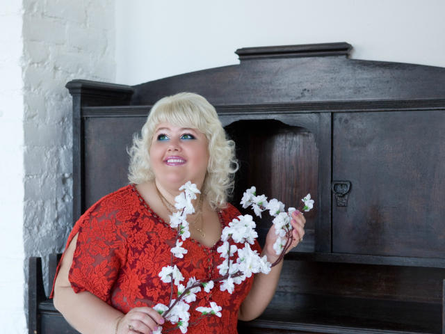 XBigBoobs with blonde curly hair, red lace dress, holding white cherry blossoms, smiling, standing in front of dark wooden cabinet, white brick wall on left.
