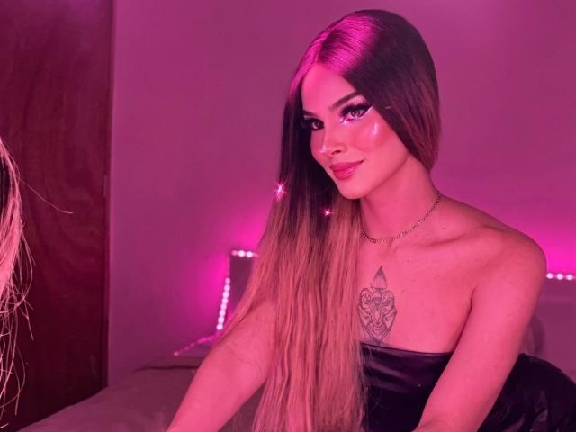 KateBmoss with long brown hair, purple eyeshadow, shiny pink lipstick, black strapless dress, heart tattoo on chest, pink neon lights, smiling, necklace, sitting on bed.