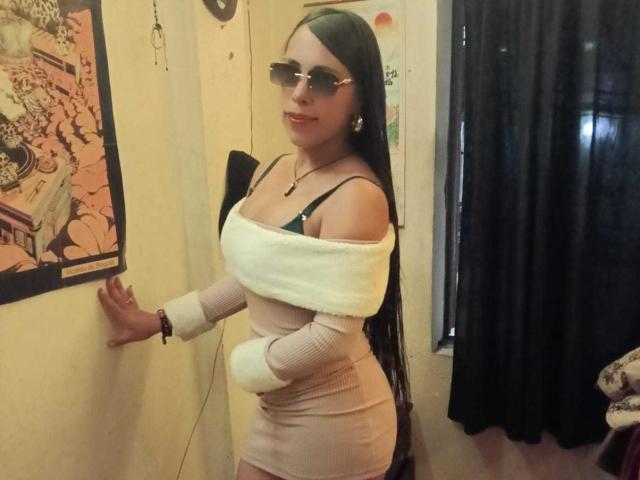 ValentinaRicci with long black hair, sunglasses, and a tight beige dress with off-shoulder white fur, standing against a wall, her big tits and small ass visible.