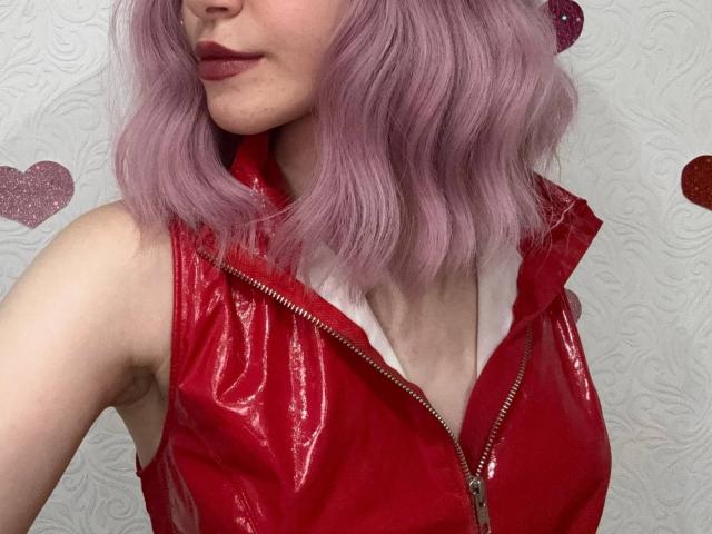 MiyaLila with pink wavy hair, wearing a red shiny zip-up vest, showing her upper body, with red lipstick and a slight smile, exposing her white shirt collar.