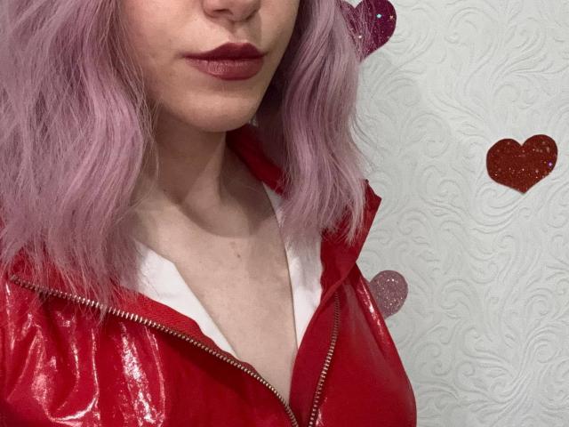 MiyaLila with purple hair, red leather jacket, white shirt, and red lipstick, showing a subtle cleavage, with glittery red hearts in the background.