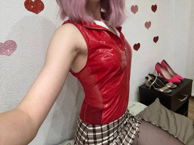 MiyaLila with pink hair, red shiny sleeveless top, plaid skirt, and pink heels, showing armpit and small tits, sitting on bed, legs apart, white wall with hearts.