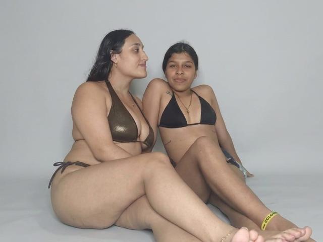 SaraAndGaby sitting on a gray floor, Sara in a brown bikini with shiny top, Gaby in a black bikini, both with medium tits, dark hair, and tan skin.