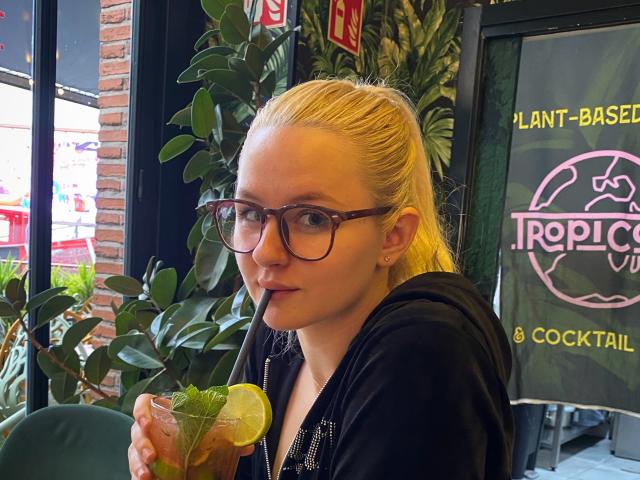Angelbysoull, con una coleta rubia, gafas, sudadera negra, tomando una bebida con una rodaja de limón, sentada en una cafetería llena de plantas. Está sorbiendo con una pajita y mirando a la cámara..