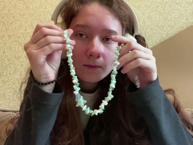 Marla Langer, con lunghi capelli castani, che tiene una collana di perline verdi e bianche, indossa delle cuffie e un cappuccio grigio scuro, e sembra leggermente infastidita. Il suo naso è leggermente rosso..